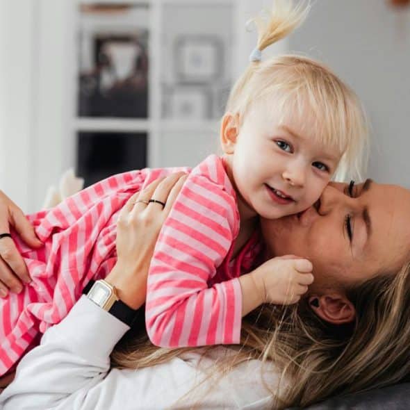 A mother lying on a couch kissing her smiling toddler in a pink striped dress.