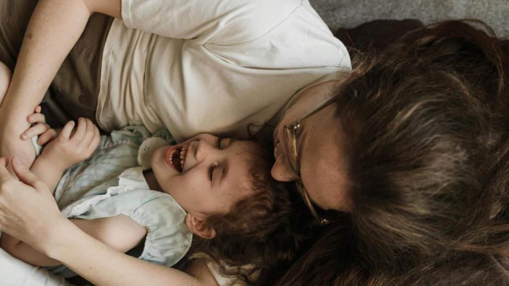 A mother lying on a bed cuddling and laughing with her young daughter.