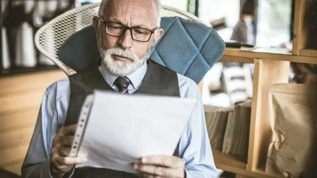 An older, well-dressed man with a beard and glasses reviews paperwork while seated indoors.