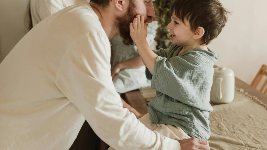 A smiling child sitting on a table touching their father’s face.