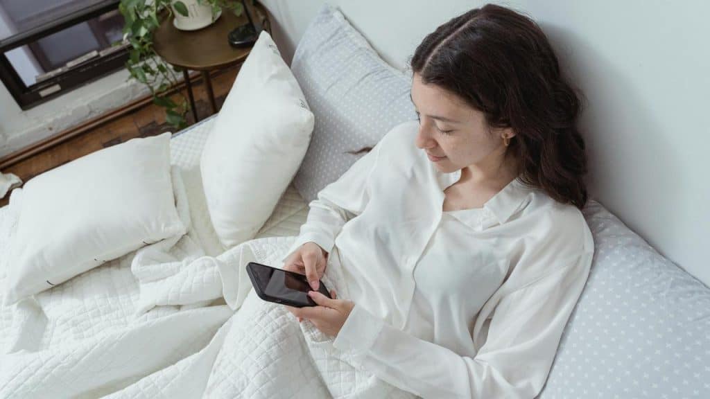 A woman sitting up in bed looking at her smartphone.