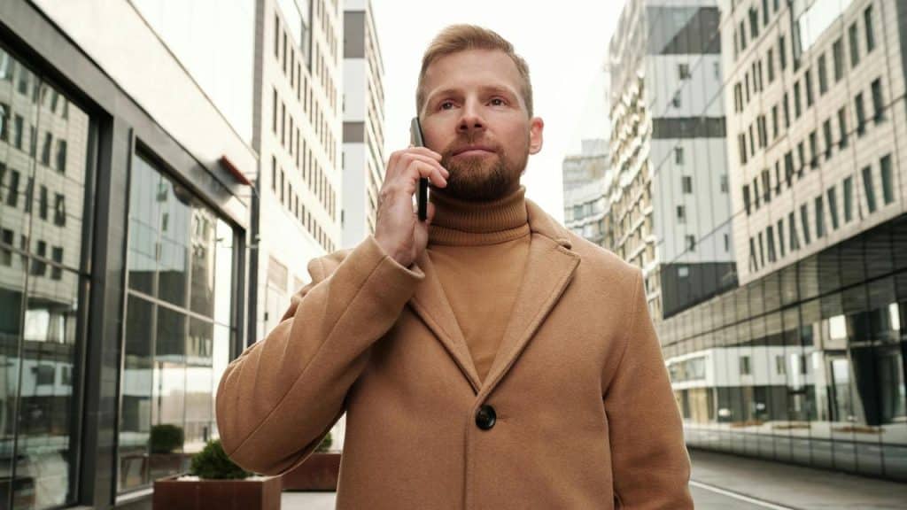 A man in a tan coat talking on his phone while standing on a city street.