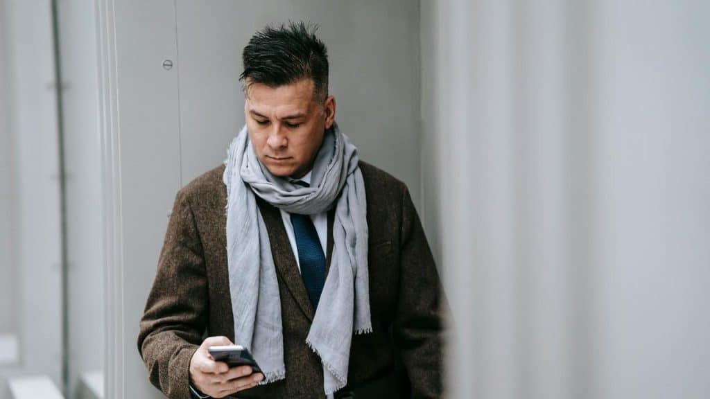 A man in a brown coat and gray scarf looking at his smartphone.