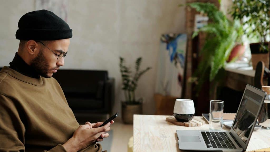A man sitting at a desk using his phone beside a laptop.