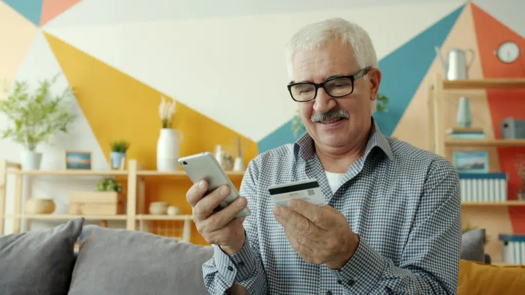 A smiling older man holds a mobile phone and a credit card in a brightly decorated room.