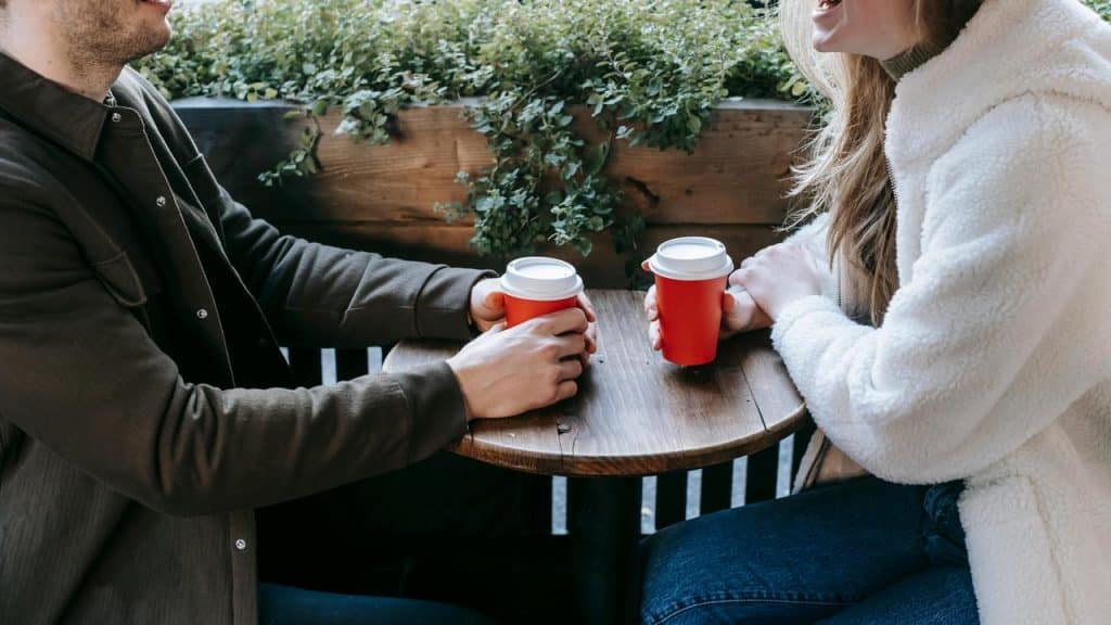 A man and a woman sitting at a small table holding red takeaway coffee cups.