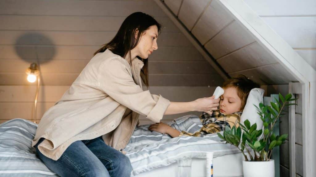 A mother checking her sick child’s temperature while he rests in bed.