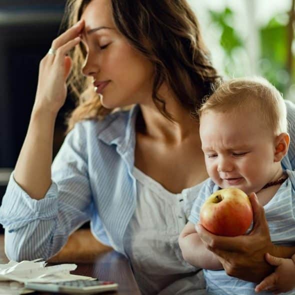 A tired mother holding a crying baby with an apple.