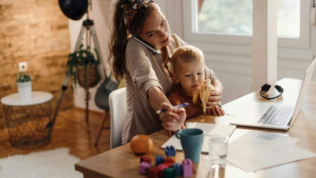 A mother working at a desk while holding her child who is eating a banana.