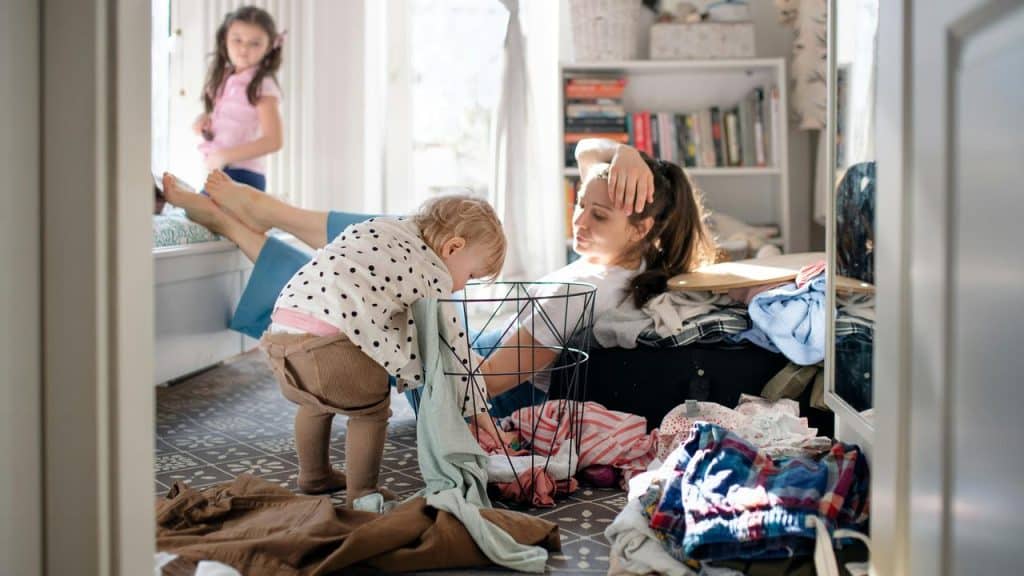 A tired mother sitting on the floor surrounded by laundry while her toddler plays nearby.