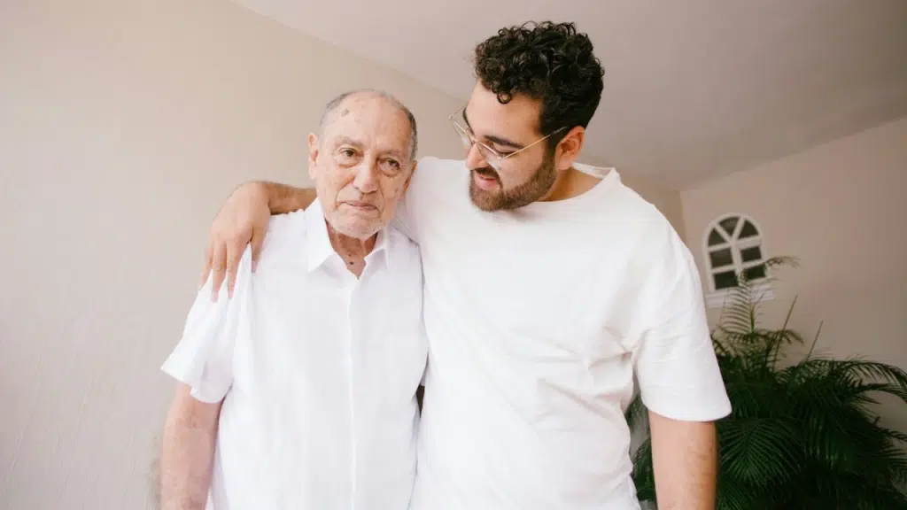 A younger man with glasses stands with his arm around an older man in white shirts.