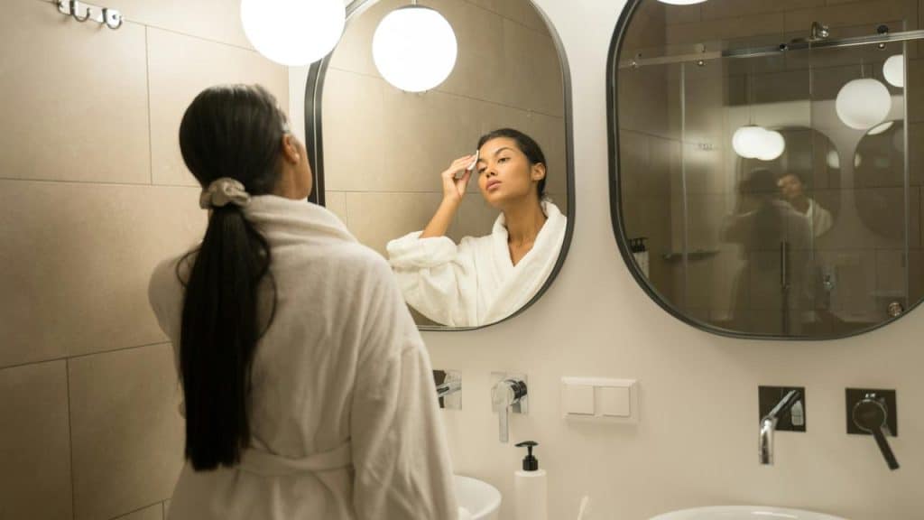 A woman in a bathrobe applying skincare in front of a bathroom mirror.