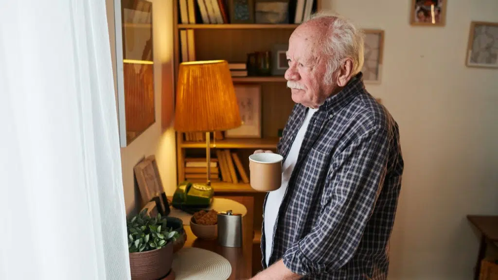 An older man in a plaid shirt stands in a living room, holding a mug and looking away.