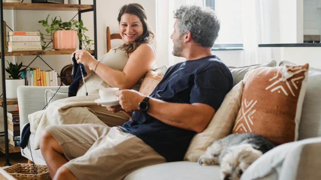 A couple sitting on a couch smiling and talking with a dog resting beside them.