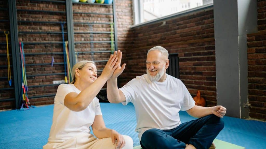 An older couple sitting on gym mats giving each other a high five.
