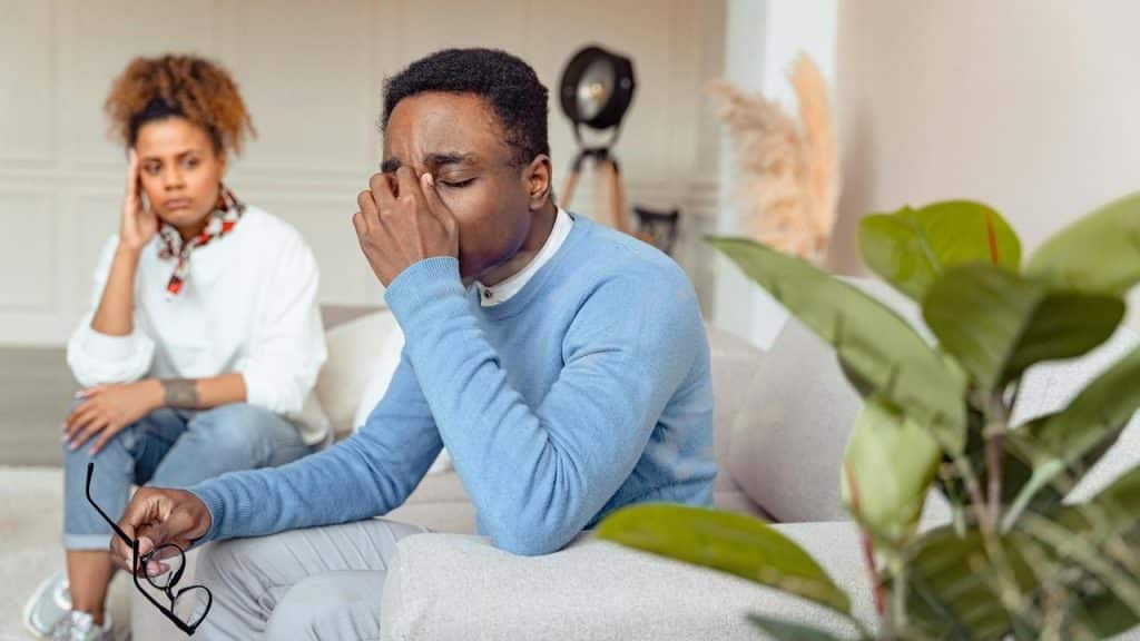 A man rubbing his eyes while holding glasses with a concerned woman behind him.