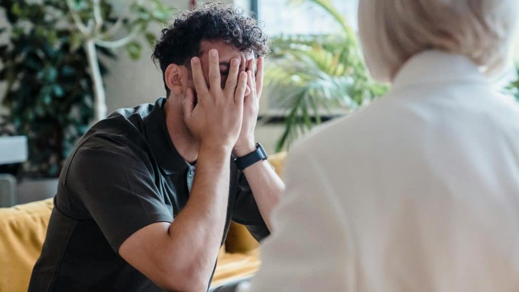 A man sitting with his face buried in his hands across from another person.