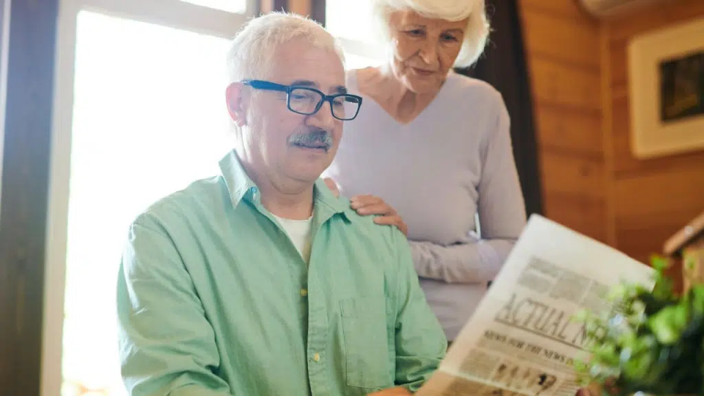 An older man with glasses reads a newspaper as an older woman stands with her hand on his shoulder.