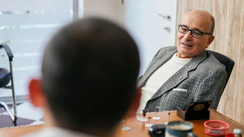An older man in a tweed jacket and glasses smiles across a desk at an unseen person.