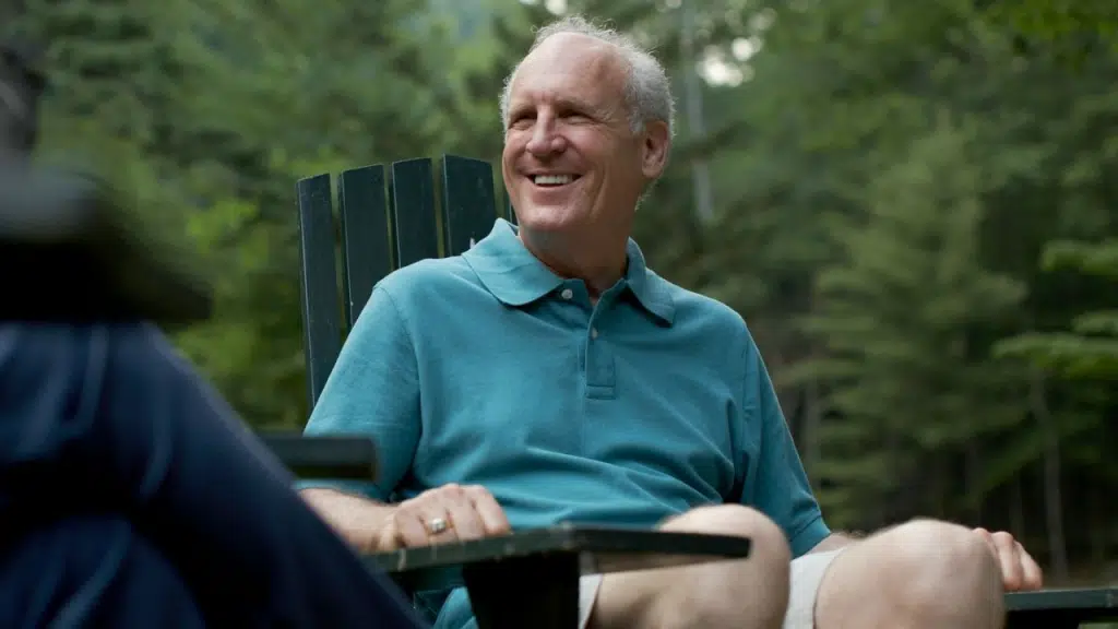 A smiling older man in a blue polo shirt sits outdoors in a wooden chair.