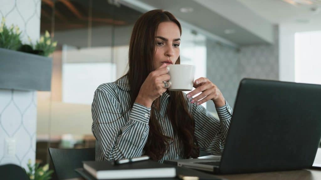A woman drinking from a cup while working on a laptop.