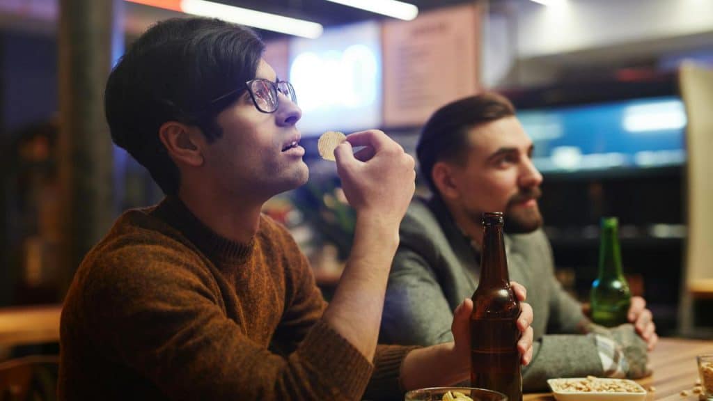 A man eating a chip at a bar next to another man with drinks.