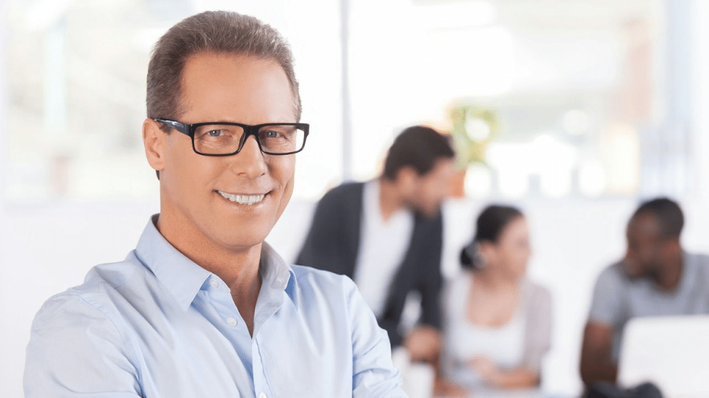 A smiling, middle-aged man in a blue shirt and glasses stands in an office setting.