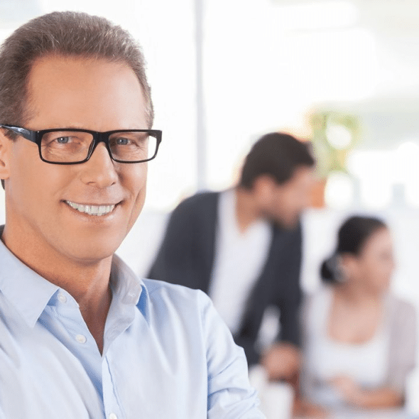 A smiling, middle-aged man in a blue shirt and glasses stands in an office setting.