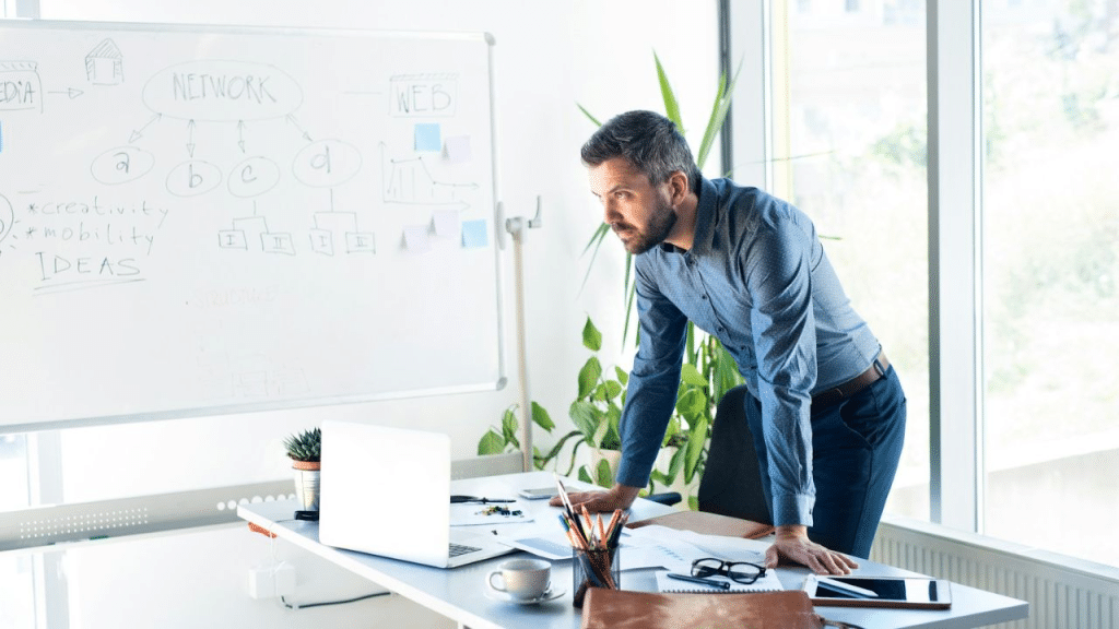 A businessman leans over his desk and looks intently at a whiteboard with diagrams.
