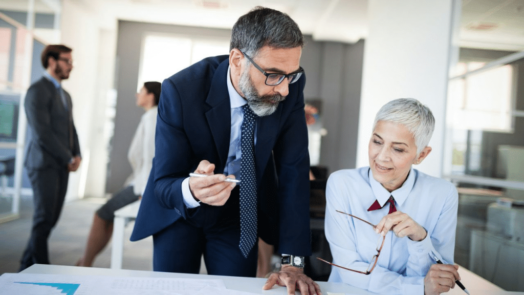A male and female business professional discuss paperwork at a desk in an office.