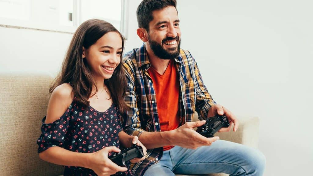 A father and daughter smiling while playing video games on the couch.