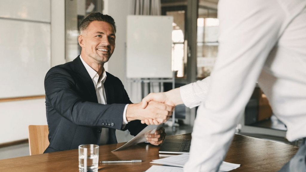 A smiling businessman in a suit shakes hands with an unseen person across a table.