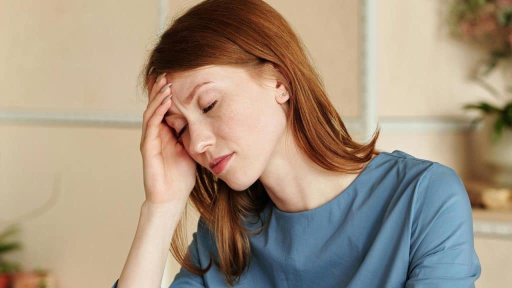 A woman in a blue top holding her head with her eyes closed.