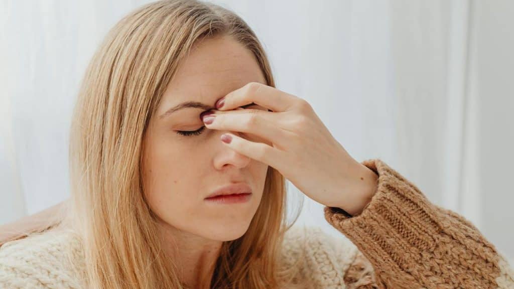 A woman in a brown sweater pinching the bridge of her nose with her eyes closed.