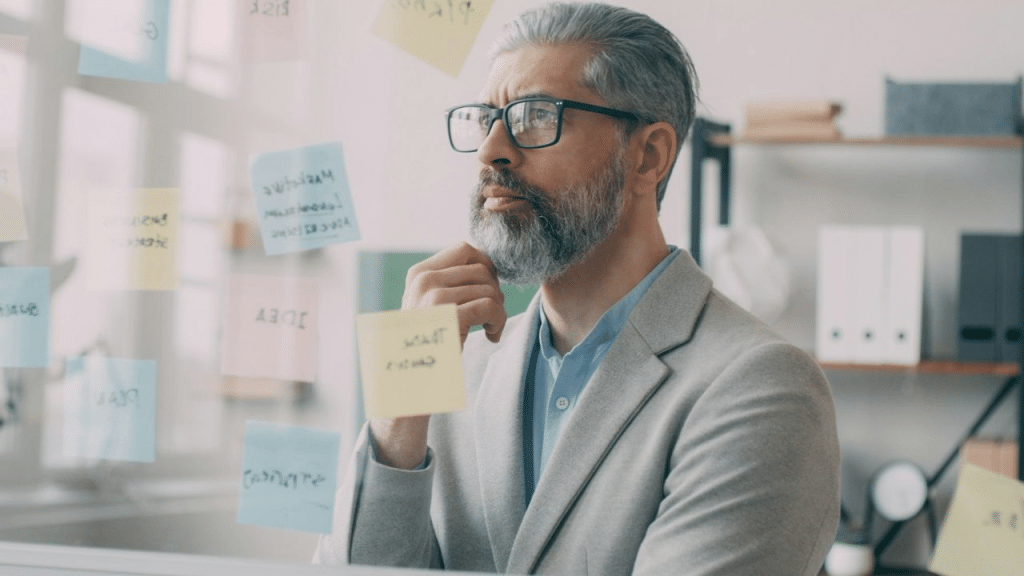 A bearded man with glasses is looking thoughtfully at a clear board covered in sticky notes.
