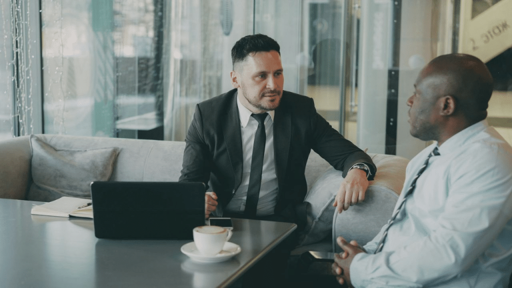 Two business professionals sit on a sofa, talking across a table with a laptop and coffee.