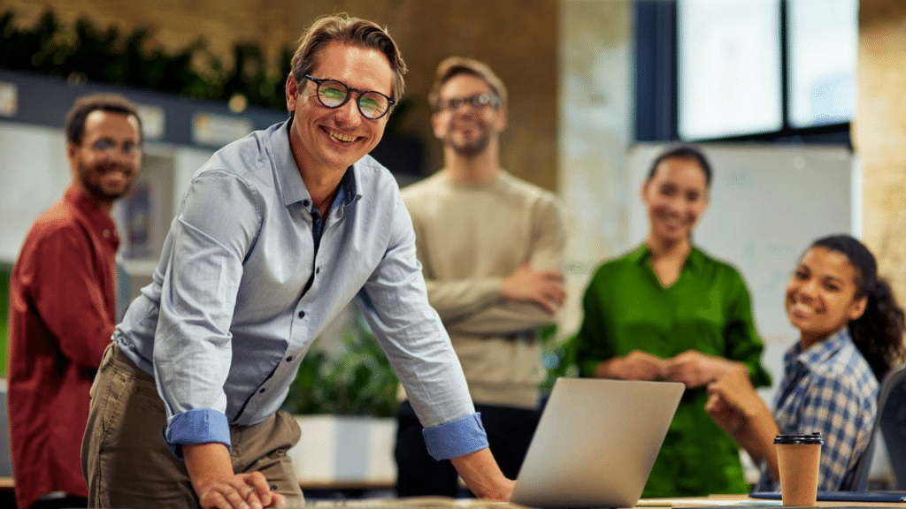 A smiling, bespectacled man leans over a laptop with four diverse colleagues behind him.