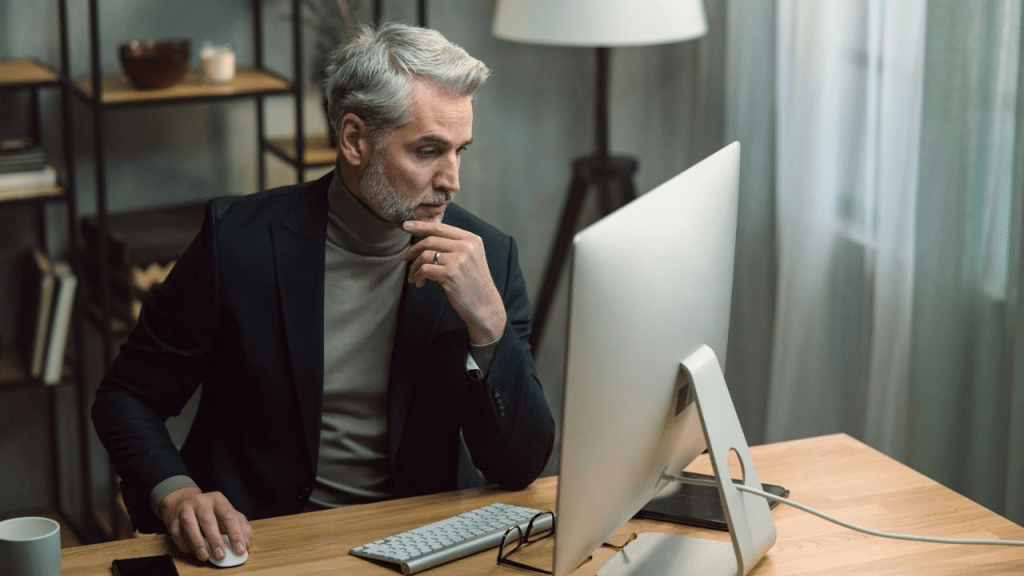 A middle-aged man in a suit jacket and turtleneck works intently at a desktop computer.