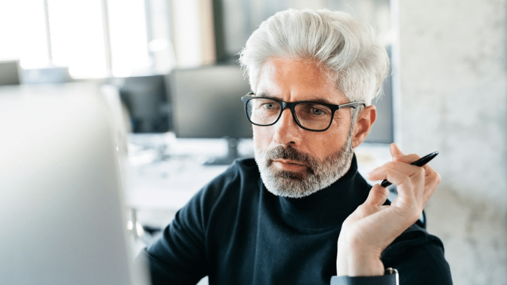 A man with white hair, a beard, and glasses looks intently at a computer screen.