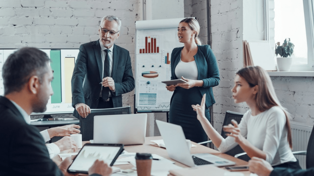 Two men and two women discuss data in a conference room with a whiteboard and monitors.