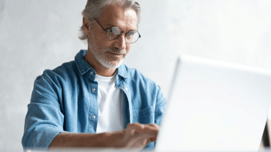 A gray-haired man in a denim shirt and round glasses works on a laptop computer.