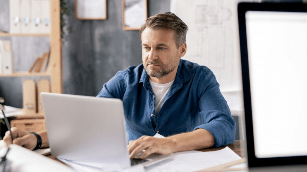 A focused, middle-aged man in a blue shirt works on a laptop at an office desk.