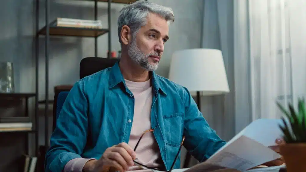 A middle-aged man with gray hair sits at a desk, looking seriously at a document.