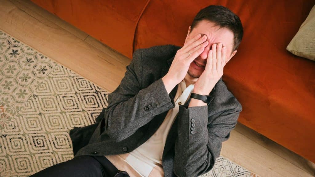 A distraught man in a blazer is crying on the floor, covering his face with his hands.