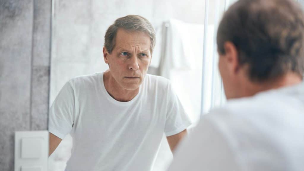 A concerned middle-aged man in a white t-shirt looks intently at his reflection in a bathroom mirror.
