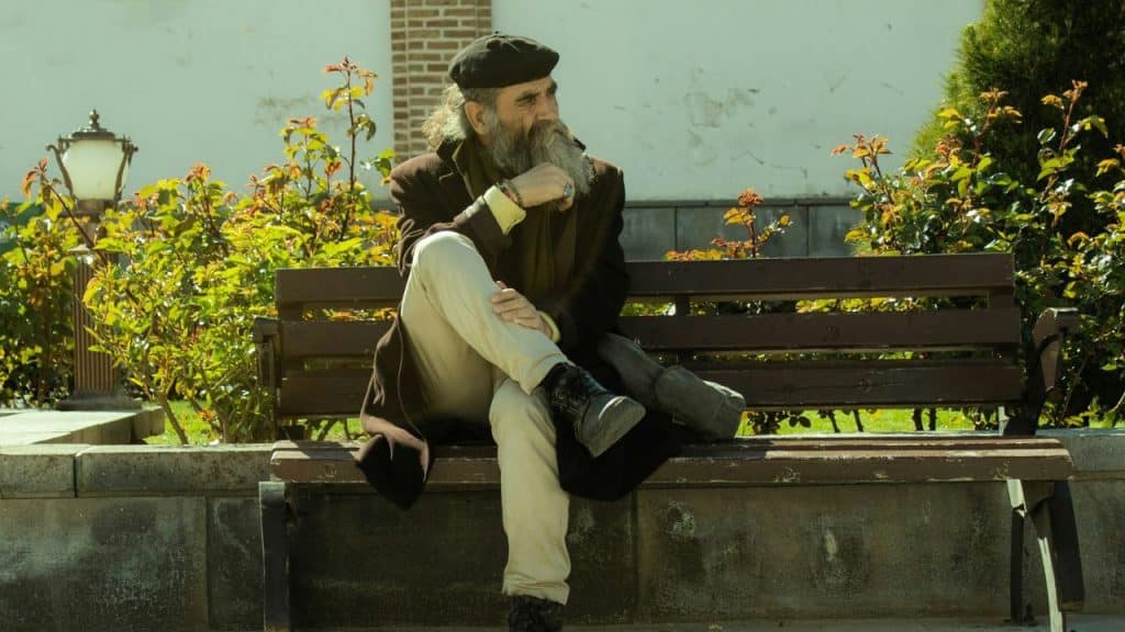 A long-bearded man in a coat and beret sits thoughtfully on a park bench.
