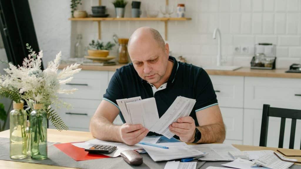 A concerned, middle-aged man sits at a kitchen table, looking over a pile of bills.