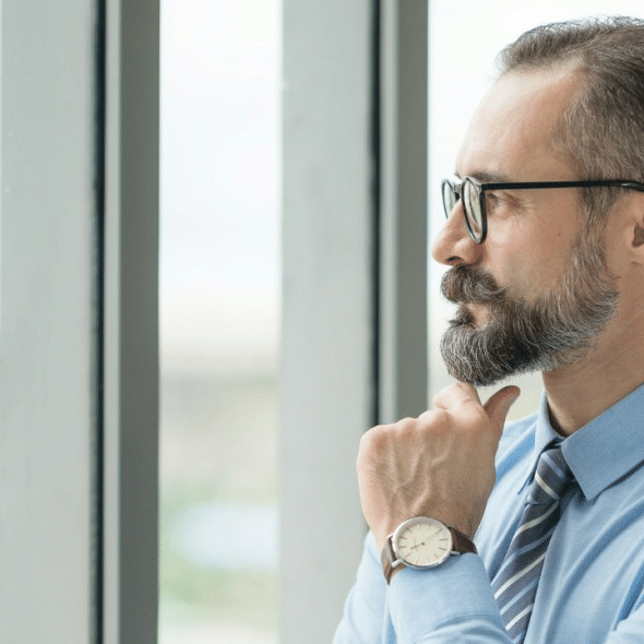 A serious man with a beard and glasses looks out a large office window, deep in thought.