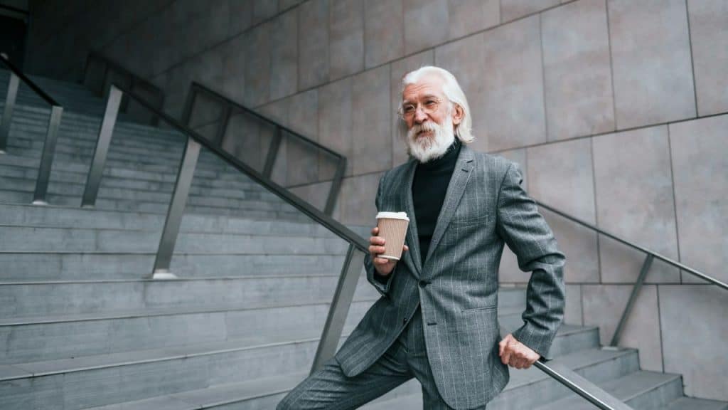 An older man in a gray suit and long beard stands on concrete stairs, holding coffee.