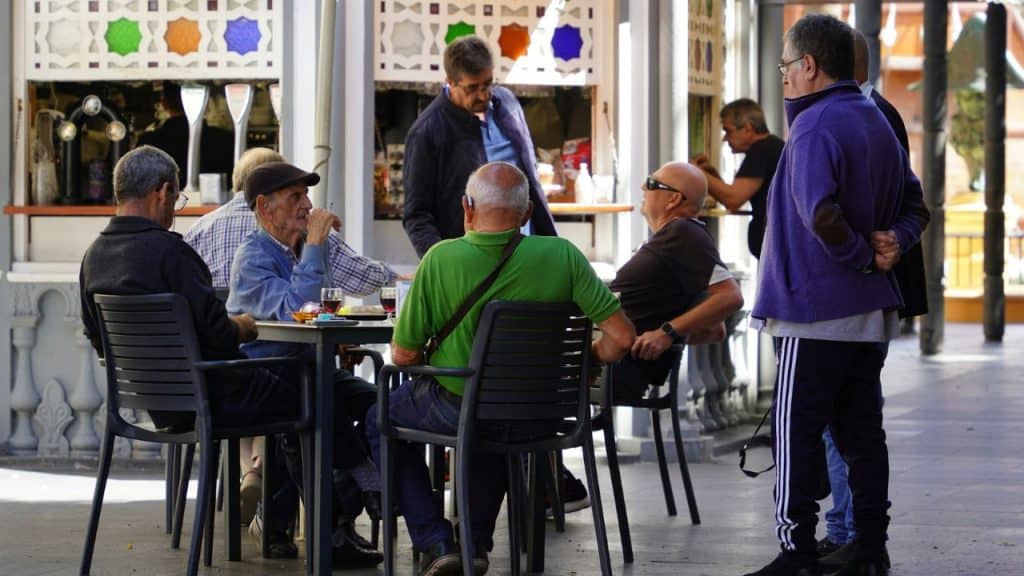 A group of older men socializes and relaxes at tables outside a cafe or bar.